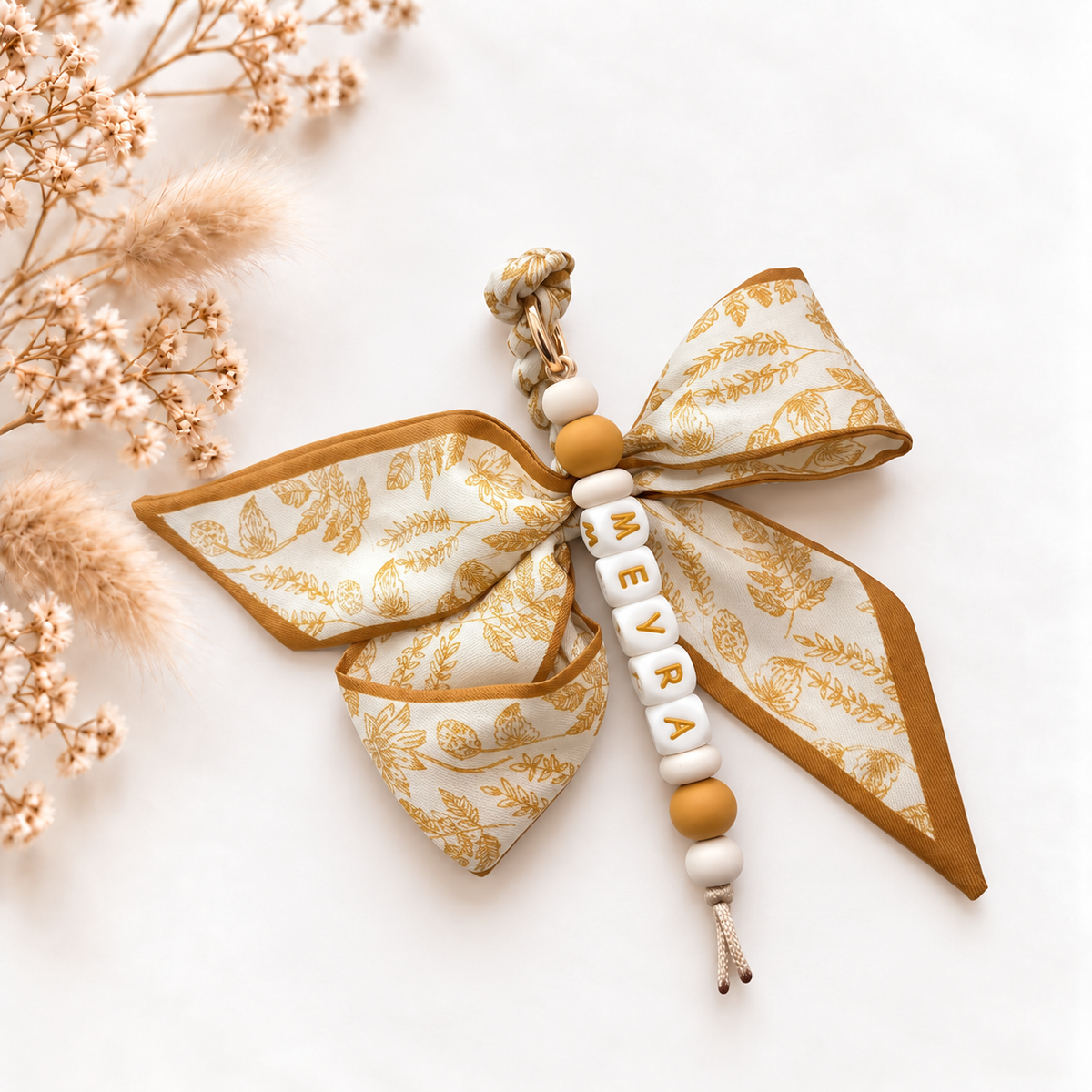 Luxe sleutelhanger en tassenhanger Golden Meadow met gepersonaliseerde naamkralen in crème- en okertinten en een grote stoffen strik met botanische bloemen- en bladerenprint.