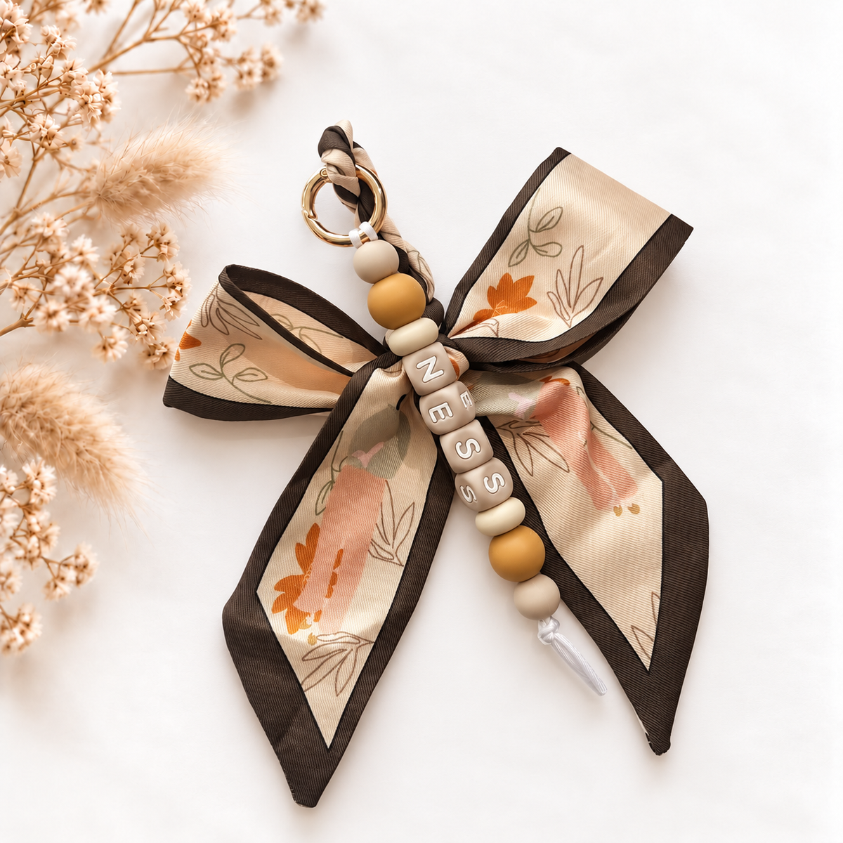 Luxe sleutelhanger en tassenhanger Amber Ribbon met gepersonaliseerde naamkralen, beige en karamelkleurige kralen en een stoffen strik met botanische print.