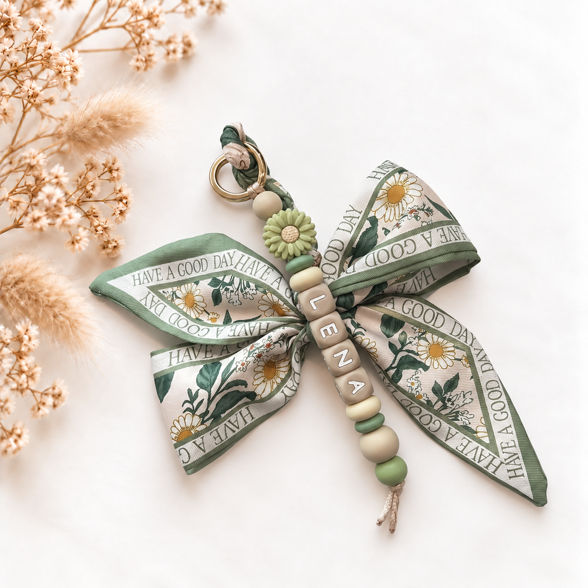 Luxe sleutelhanger en tassenhanger Green Daisy met gepersonaliseerde naamkralen, groene en beige kralen, een decoratieve bloem en een stoffen strik met botanische bloemenprint.