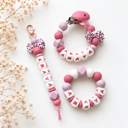 Gepersonaliseerde kraamcadeauset Floral Bow met roze speenkoord, sleutelhanger en bijtring met naam, versierd met strikjes en kralen in roze, lila en wit.