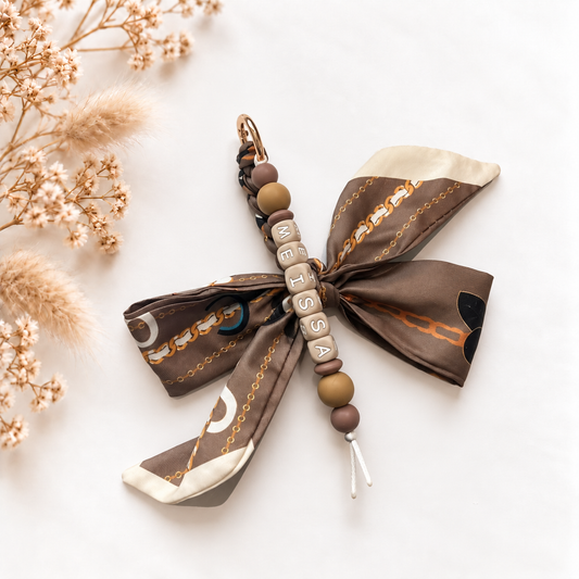 Luxe sleutelhanger en tassenhanger Mocha Chain met gepersonaliseerde naamkralen in bruine en karamelkleuren en een grote stoffen strik met beige en bruine kettingprint.